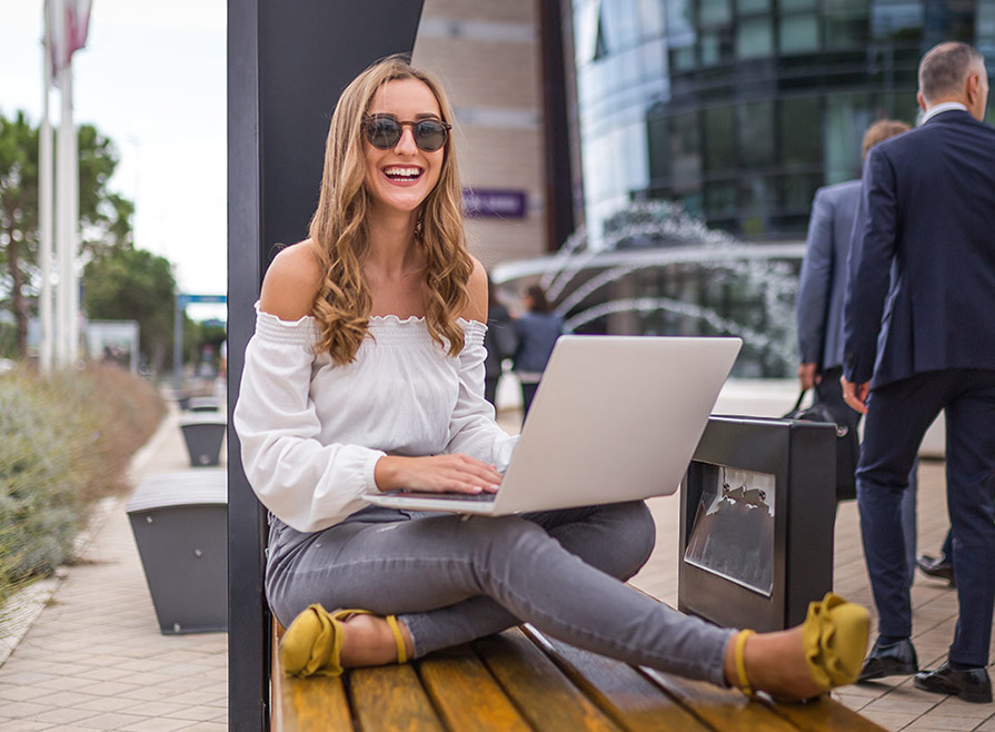 Smiling stylish young woman wearing yellow shoes sitting on yellow bench using laptop depicting browsing early career resume packages